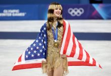 USA figure skater Alysa Liu smiles wearing her gold medal with a US flag draped over her shoulders on the 2026 Olympic podium.