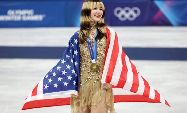 USA figure skater Alysa Liu smiles wearing her gold medal with a US flag draped over her shoulders on the 2026 Olympic podium.